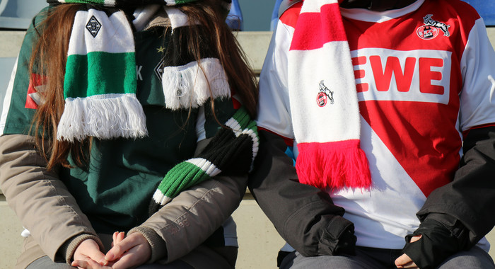 Ein Gladbach-Fan und ein Köln-Fan sitzen in Fankleidung nebeneinander