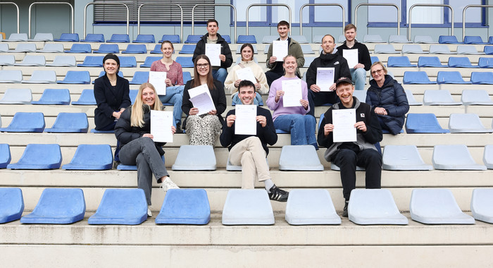 mehrere Personen sitzen auf Besucher*innenrängen eines Stadions und halten Urkunden hoch