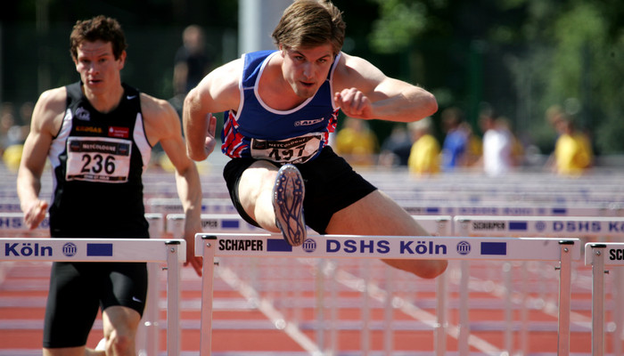 Ein Sportler läuft über eine Hürde, auf der DSHS Köln steht.