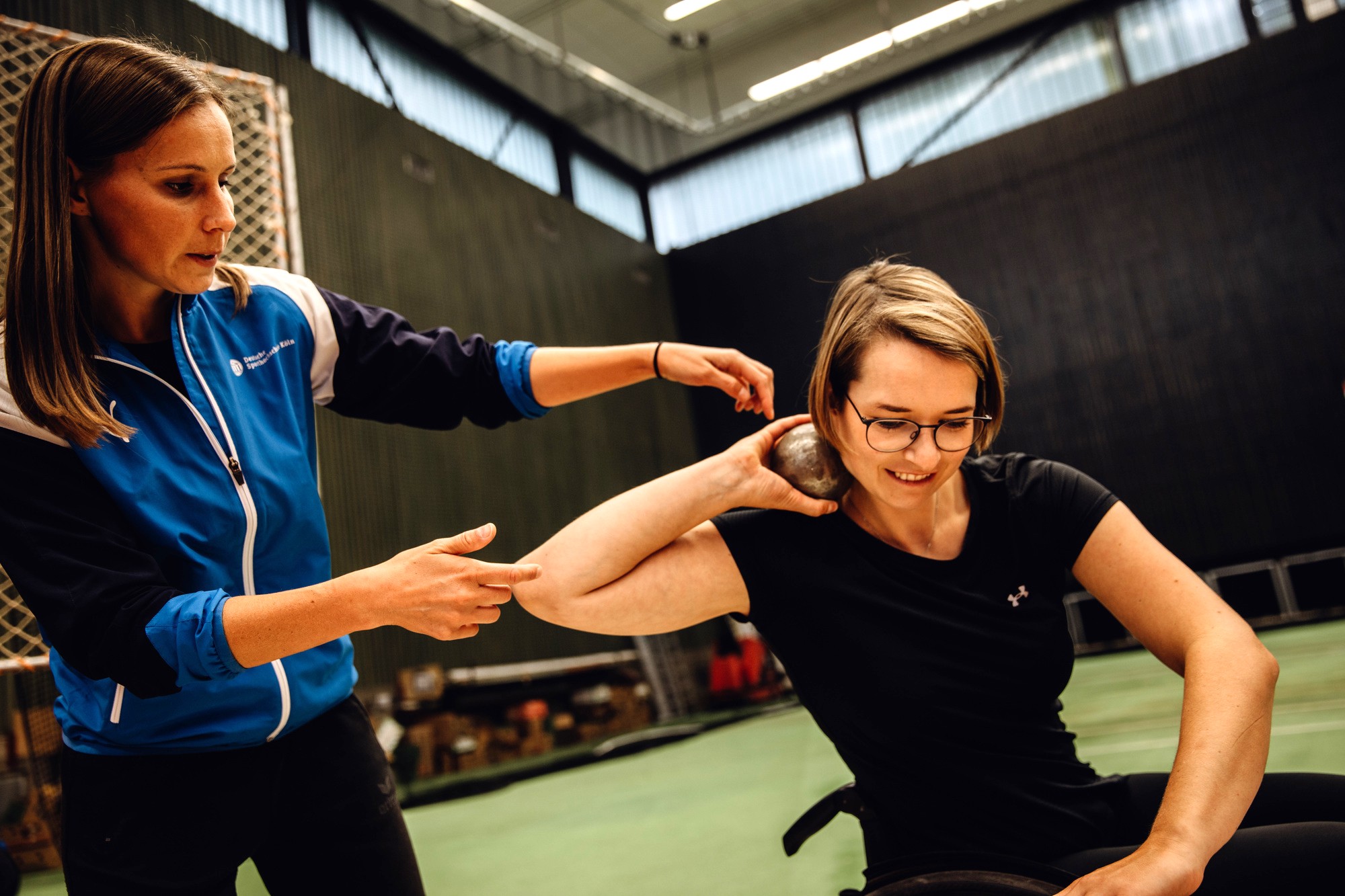 Eine Studentin im Rollstuhl befindet sich im Kugelstoßring. Die Kugel ist seitlich am Hals platziert, der Ellenbogen ist dabei seitlich hinter der Kugel. Die Dozentin steht links neben dem Rollstuhl. 
