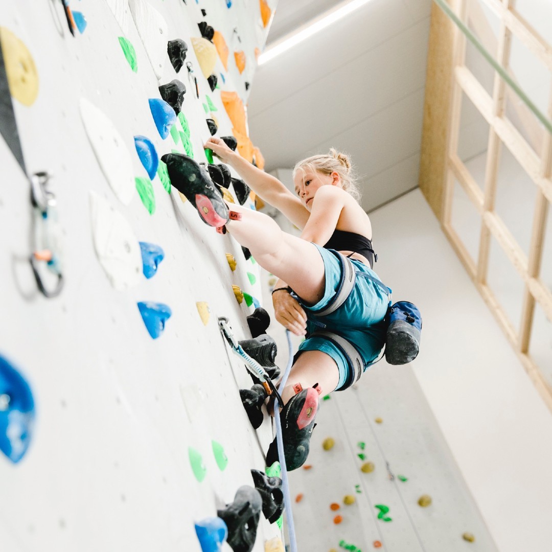 Frau in Sportbekleidung klettert an einer Kletterwand mit bunten Griffen in einer Halle
