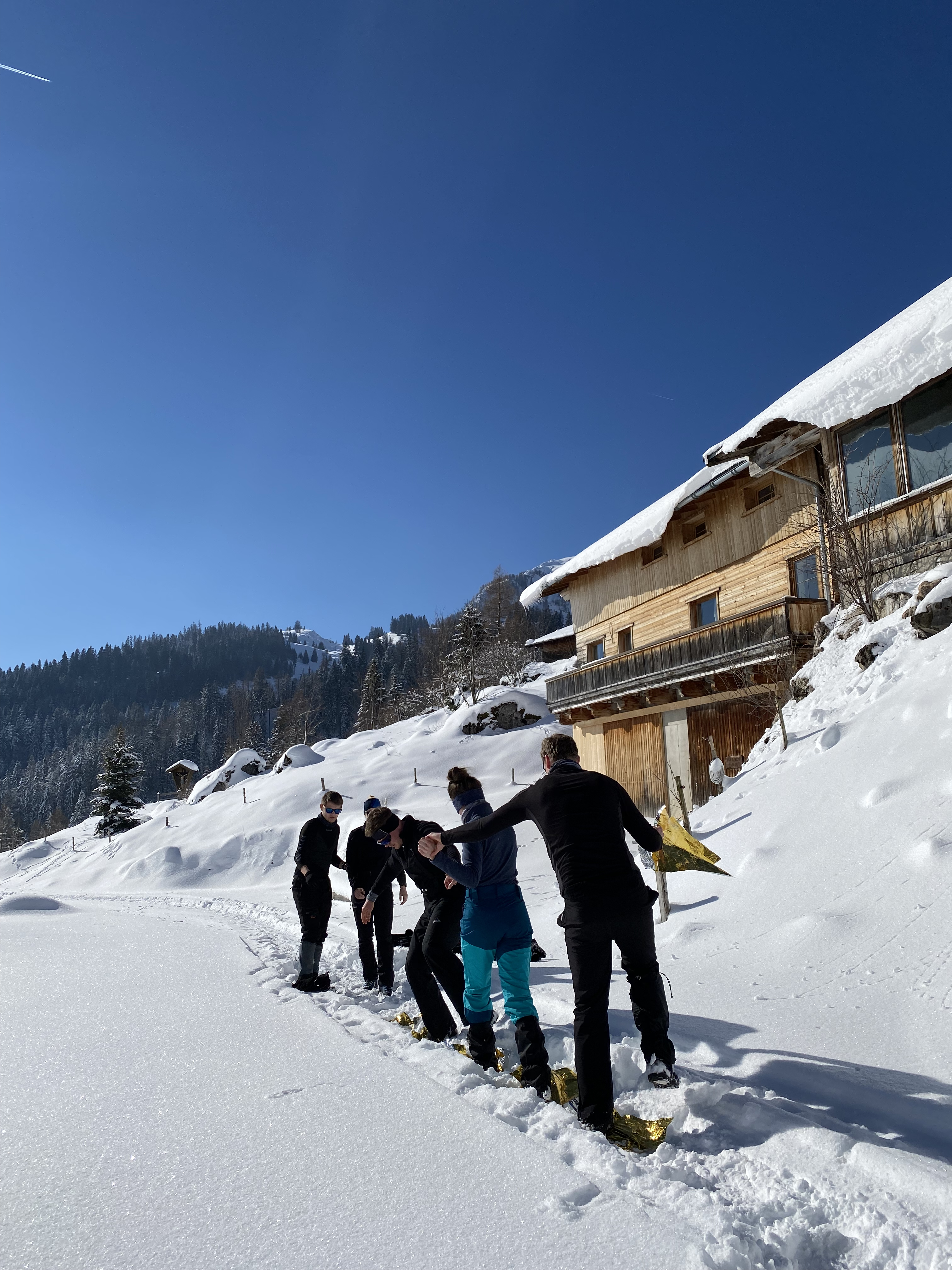 Gruppe von Personen mit Schneeschuhen läuft in einer Reihe auf einem verschneiten Weg neben einer Holzhütte unter klarem blauem Himmel
