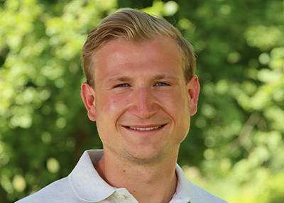 Porträt-Foto von Felix Eiting. Felix ist ein blonder Mann und trägt ein weißes Hemd. Im Hintergrund sind Bäume zu sehen. Er lächelt in die Kamera.

