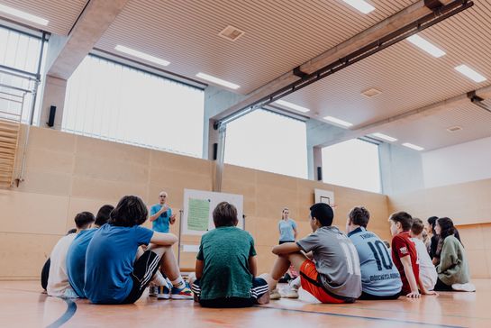 Kinder sitzen in einer Turnhalle im Kreis und eine Lehrkraft erklärt etwas an einem Board.