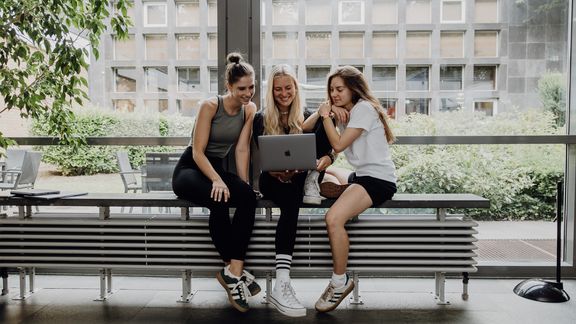 Drei Studentinnen arbeiten gemeinsam an einem Laptop im Campusgebäude.
