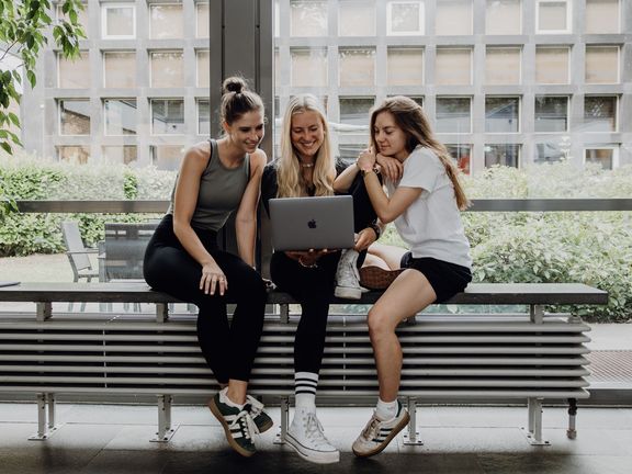 Drei Studentinnen arbeiten gemeinsam an einem Laptop im Campusgebäude.
