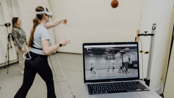 Forschende Person analysiert am Laptop während Probandin mit VR-Brille einen Basketball im leeren Raum wirft
