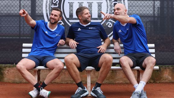 Drei Männer in Sportkleidung sitzen auf einer weißen Bank auf einem Tennisplatz und zeigen in verschiedene Richtungen. Im Hintergrund ist das Logo der Sporthochschule zu sehen. 
