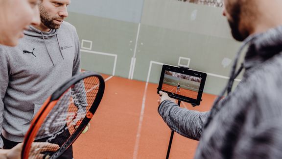Foto von drei Personen die eine Tennisaufnahme auf einem Tennisplatz betrachten
