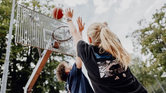 Foto von einem Basketballwurf in Richtung Korb von unten, den ein Mann versucht zu verteidigen
