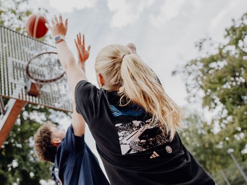 Foto von einem Basketballwurf in Richtung Korb von unten, den ein Mann versucht zu verteidigen
