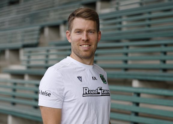 Porträtfoto von Fabian Hellberg. Er steht leicht seitlich in einem weißen Sportshirt vor einer Tribüne mit Bänken.
