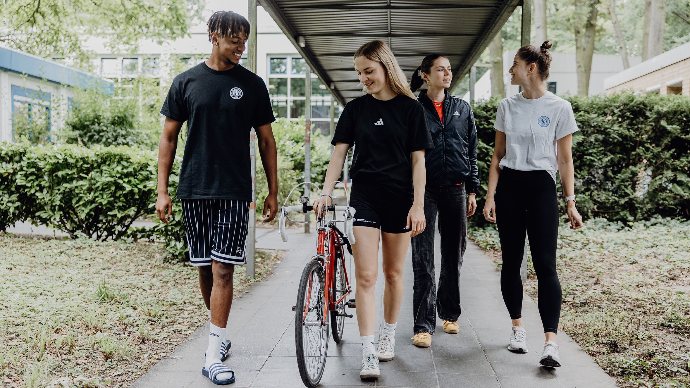 Vier Studierende gehen mit einem Rennrad über den Campus der Deutschen Sporthochschule
