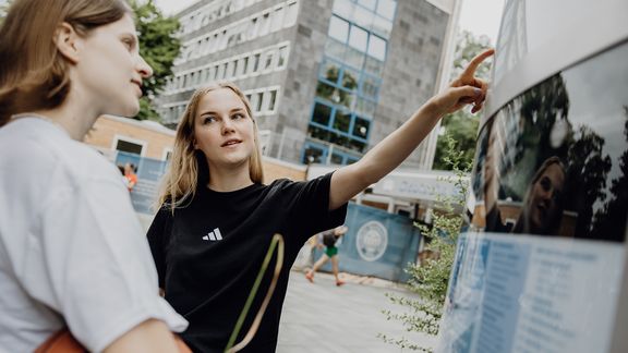 Zwei Studierende zeigen auf den Wegweiser im Eingangsbereich der Deutschen Sporthochschule
