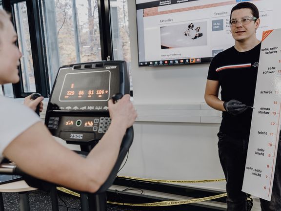 Foto von forschender Person der einer auf dem Ergometer sitzenden Probandin eine Skala zeigt
