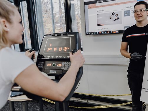 Foto von forschender Person der einer auf dem Ergometer sitzenden Probandin eine Skala zeigt

