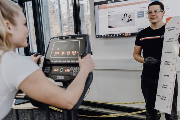 Foto von forschender Person der einer auf dem Ergometer sitzenden Probandin eine Skala zeigt

