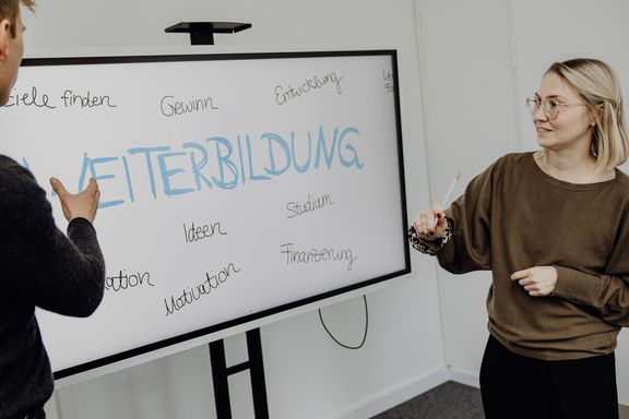 Foto von zwei Personen die vor Whiteboard mit dem Schriftzug "Weiterbildung" stehen
