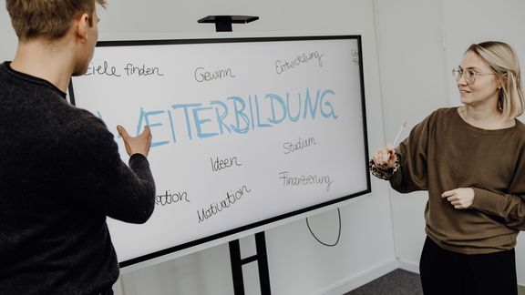 Foto von zwei Personen die vor Whiteboard mit dem Schriftzug "Weiterbildung" stehen
