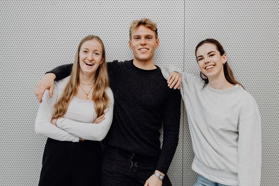 Drei Studierende stehen Arm in Arm nebeneinander