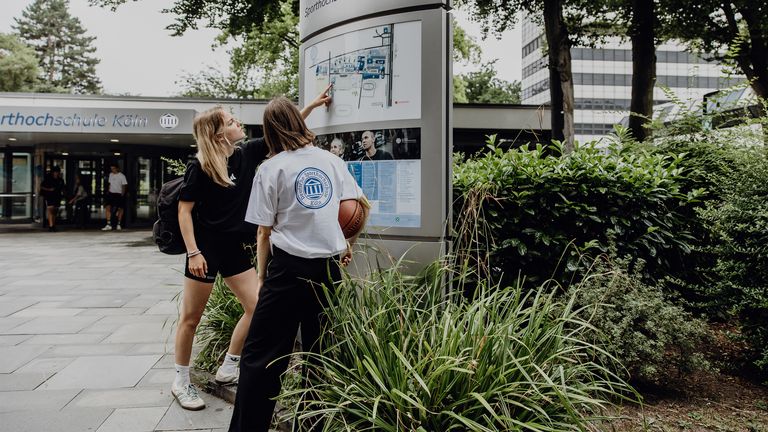 Zwei Studierende betrachten den Wegweiser im Eingangsbereich der Deutschen Sporthochschule
