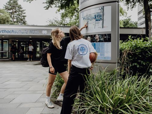 Zwei Studierende betrachten den Wegweiser im Eingangsbereich der Deutschen Sporthochschule
