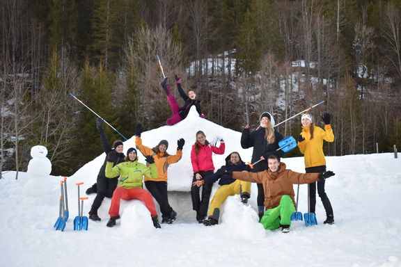 Gruppe von neun Personen vor einem Iglu aus Schnee mit Schneeschaufeln in der Hand und einem Schneemann im Hintergrund
