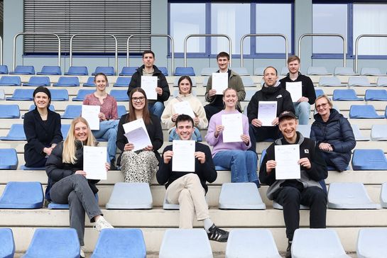 Absolvent*innen sitzen mit ihren Zertifikaten auf der Tribüne des LAA-Stadions