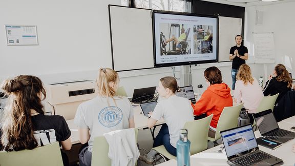 Studierende sitzen im Seminar und hören Dozierendem zu
