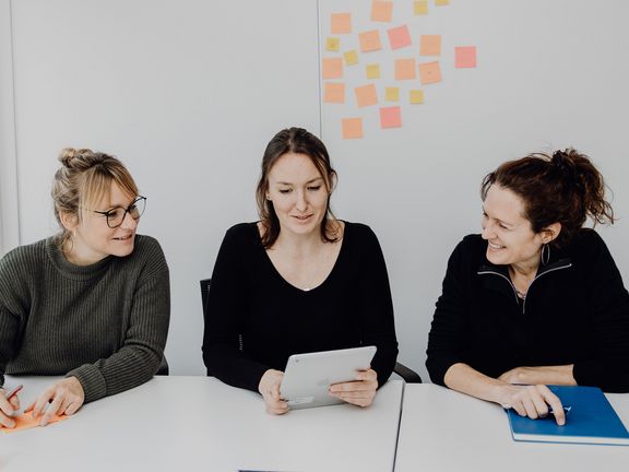 Foto von drei Frauen die am Tisch sitzen und auf ein Tablet in der Mitte blicken
