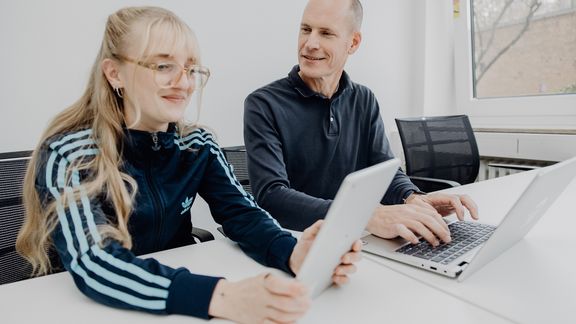 Foto von zwei Personen die an einem Tisch sitzen und mit Laptop und Tablet arbeiten
