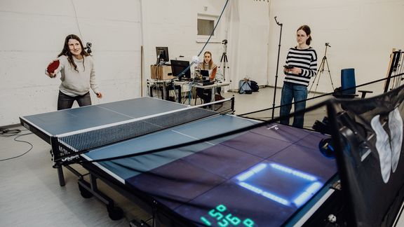 Probandin führt Tischtennisaufschlag auf einer Platte in Richtung eines leuchtenden Quadrats auf der anderen Seite aus
