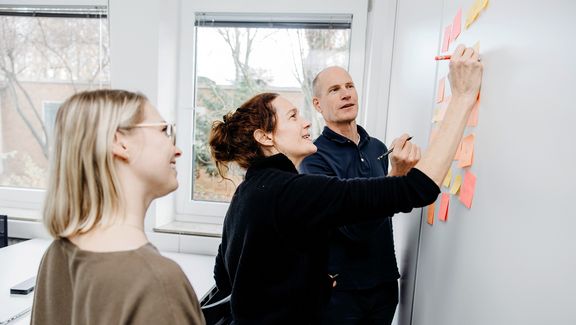 Drei Personen beschriften kleine, klebende Zettel auf einem Whiteboard
