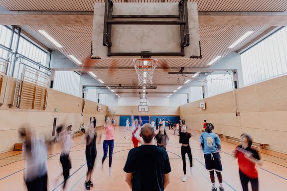 Lehrkraft schaut zu einen Basketballkorb auf den gleichzeitig mehrere Kinder einen Basketball werfen
