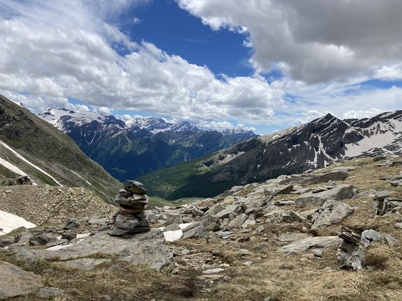 Felsiger Bergpfad mit zwei Steintürmen und schneebedeckten Gipfeln im Hintergrund unter bewölktem Himmel
