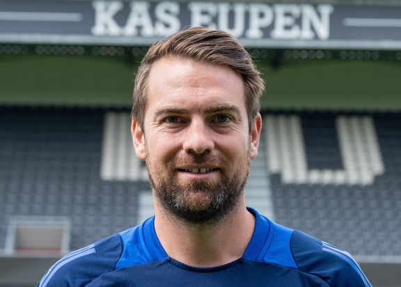 Porträtfoto von Nicolas Collubry. Er trägt ein blaues Trikot und steht vor der grauen Tribüne des Stadions vom KAS Eupen

