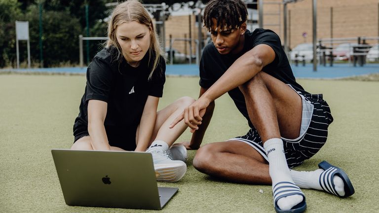 Zwei Studierende sitzen auf einer Sportfläche im Freien und arbeiten gemeinsam an einem Laptop.
