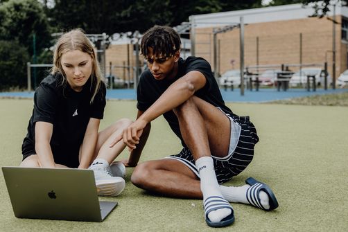 Zwei Studierende sitzen auf einer Sportfläche im Freien und arbeiten gemeinsam an einem Laptop.
