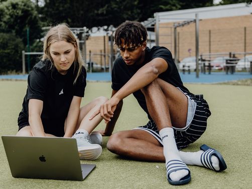 Zwei Studierende sitzen auf einer Sportfläche im Freien und arbeiten gemeinsam an einem Laptop.
