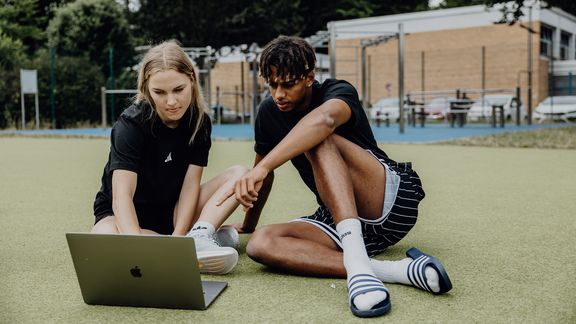 Zwei Studierende sitzen auf einer Sportfläche im Freien und arbeiten gemeinsam an einem Laptop.
