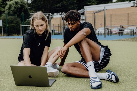 Zwei Studierende sitzen auf einer Sportfläche im Freien und arbeiten gemeinsam an einem Laptop.
