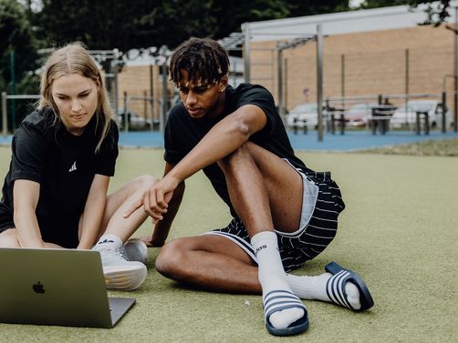 Zwei Studierende sitzen auf einer Sportfläche im Freien und arbeiten gemeinsam an einem Laptop.
