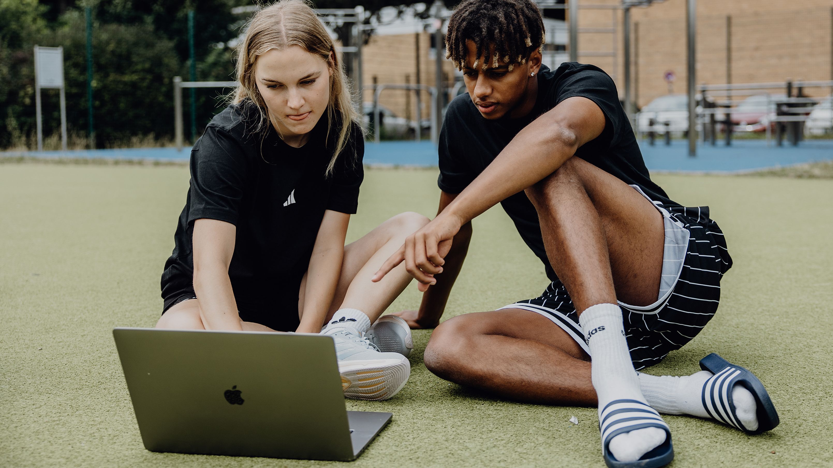 Zwei Studierende sitzen auf einer Sportfläche im Freien und arbeiten gemeinsam an einem Laptop.
