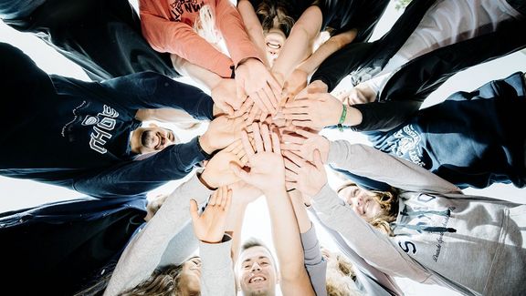 Im Kreis stehende Studierende legen die Hände in der Mitte übereinander
