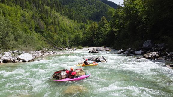 Drei Personen in bunten Kajaks paddeln auf einem Wildfluss, der von Felsen durchsetzt ist. Das Wasser ist klar und schäumt an den Steinen. Dichte grüne Wälder bedecken die steilen Hänge zu beiden Seiten des Flusses. Im Hintergrund ragen bewaldete Berge auf. Die Szene wirkt naturnah und dynamisch.
