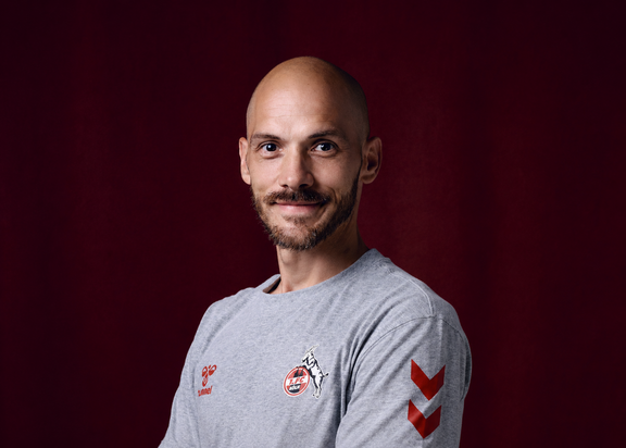 Porträt von Manuel Hartmann. Er trägt ein graues 1. FC Köln-T-Shirt und steht vor einem weinroten Hintergrund
