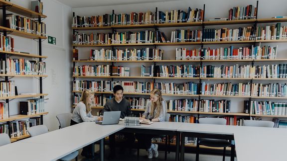 Drei Studierende sitzen mit Büchern und Laptop vor einem großen Bücherregal
