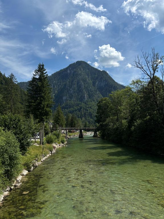Ein klarer Fluss fließt durch eine bewaldete Landschaft. Im Hintergrund erhebt sich ein bewaldeter Berg unter einem blauen Himmel mit einzelnen weißen Wolken. Eine schmale Brücke überspannt den Fluss. Am Ufer wachsen dichte Bäume und Sträucher.
