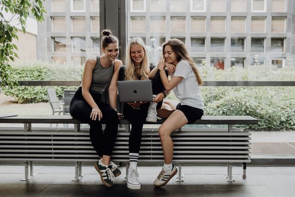 Drei Studentinnen arbeiten gemeinsam an einem Laptop im Campusgebäude.
