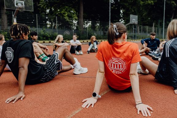 Studierende der Deutschen Sporthochschule Köln sitzen im Kreis auf dem Basketball-Court im Freien.
